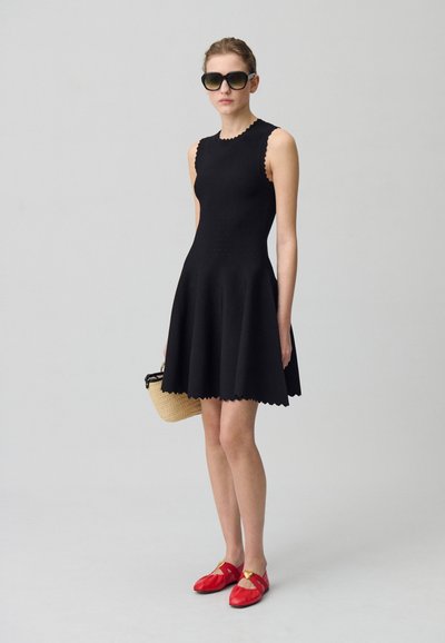 Robe noire sans manches avec un ourlet festonné, associée à des chaussures rouges à enfiler et un petit sac tissé. Tissu solide, silhouette ajustée et texture lisse.