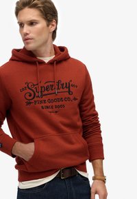 Sudadera de color óxido con un bolsillo frontal, capucha ajustable con cordón y marca impresa. Cuenta con puños y bajo de canalé, fabricada con tejido suave.