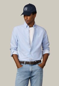 Muškarac nosi tamnoplavu kapu, svijetloplavu košulju s gumbima preko bijele majice, svijetloplave traperice i tamnosmeđi pleteni remen, ruke u džepovima.