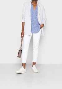 Person med lys lilla blazer, blå blomsterbluse, hvide cropped bukser og hvide sneakers, der holder en beige håndtaske ved siden af.