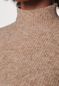 Pull en tricot beige avec un col côtelé haut. Tissu doux et texturé avec un motif subtil, mettant en avant la chaleur et le confort.