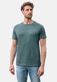 T-shirt verde a maniche corte in tessuto morbido, con scollatura rotonda e un piccolo logo sul petto. Indossato con jeans blu.