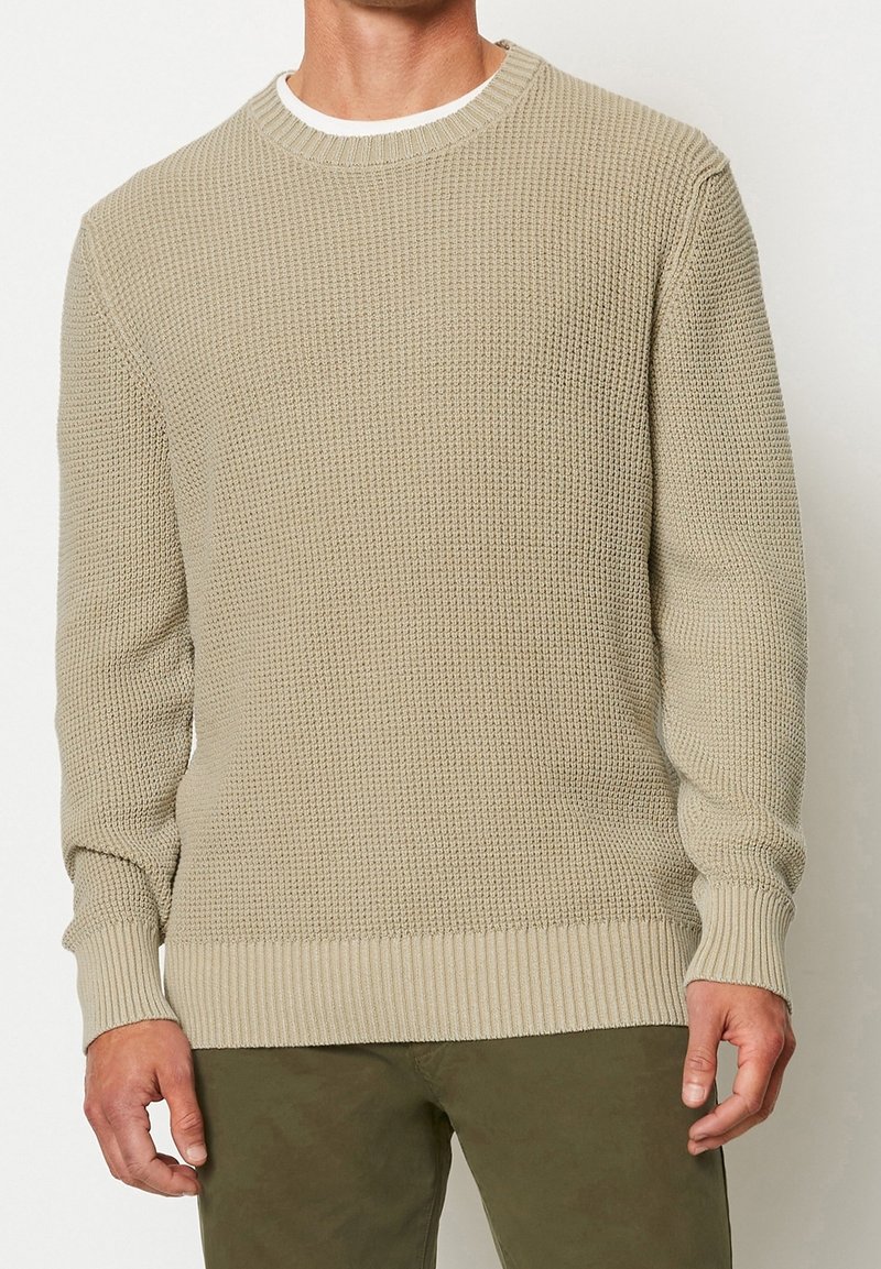 Homme portant un pull en maille gaufrée beige sur une chemise blanche et un pantalon vert olive, debout devant un fond clair uni.