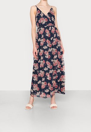 Frau trägt ein ärmelloses, marineblaues Maxikleid mit pinkem Blumenmuster und weißen Sandalen mit Absatz, steht vor einem einfarbigen Hintergrund.