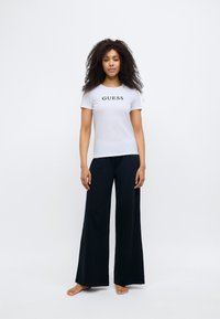 White short-sleeve t-shirt with black "GUESS" logo, paired with black wide-leg pants. Smooth fabric, relaxed fit, minimal design.