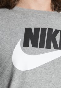 Halliksvoo hall puuvillane T-särk, mille esiküljel on suur must "NIKE" logo ja valge suti kujundus, pehme ja tekstuuriga viimistlusega.