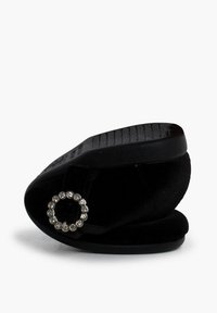 Zapatilla de terciopelo negro sin cordones, con un acento de joya redonda y suela texturada negra. Presenta un perfil bajo y un diseño de punta redonda.