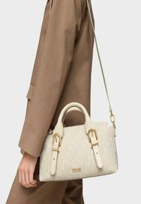 Bolso de mano blanco con patrón en relieve, herrajes en tono dorado, asas superiores dobles y una correa de hombro desmontable, llevado por una modelo.