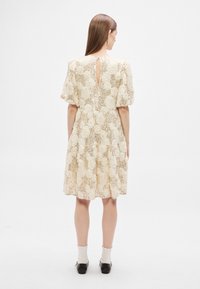 Φόρεμα σε κρεμ απόχρωση με υφασμάτινο floral ανάγλυφο μοτίβο, κοντά puff μανίκια και λεπτομέρεια πίσω με άνοιγμα, συνδυασμένο με λευκές κάλτσες και μαύρα παπούτσια.