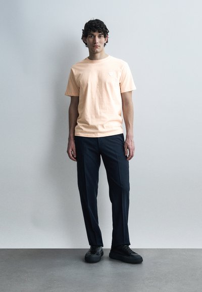 Νέος άνδρας με σγουρά μαλλιά, ντυμένος με ανοιχτό ροδακινί μπλουζάκι, μαύρο παντελόνι και μαύρα αθλητικά παπούτσια, στέκεται μπροστά σε ένα απλό ανοιχτό γκρι τοίχο.