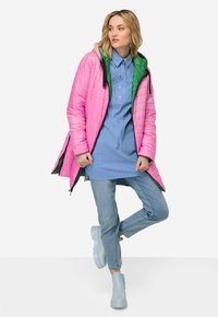 Chaqueta acolchada rosa con un forro interior verde, llevada sobre una camisa de rayas azul y unos vaqueros de denim claros. El atuendo incluye botas gruesas de color azul claro.