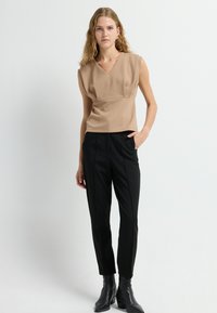 Beige mouwloos top met een V-hals en gedrapeerd ontwerp, gecombineerd met op maat gemaakte zwarte broek met verticale plooien en zijzakken. Zwarte enkellaarsjes.