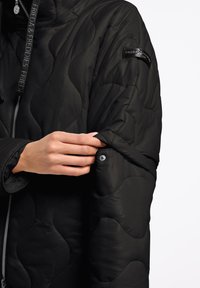 Wypełniona czarna kurtka z pikowanym wzorem. Posiada zamek błyskawiczny, naszywkę z logo na rękawie oraz zapięcie na nap na mankiecie.