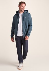 Teal zip-up hoodie with a front pocket, drawstring hood, and logo. Worn over a white t-shirt, paired with dark jeans and white sneakers.