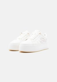 Baskets à plateforme blanches avec design à lacets et broderie florale sur les côtés, présentées sur un fond blanc uni.