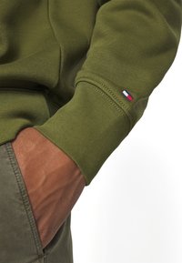 Personne portant une chemise à manches longues vert olive avec un petit logo rouge, blanc et bleu sur le poignet, la main partiellement dans la poche d'un pantalon vert olive.