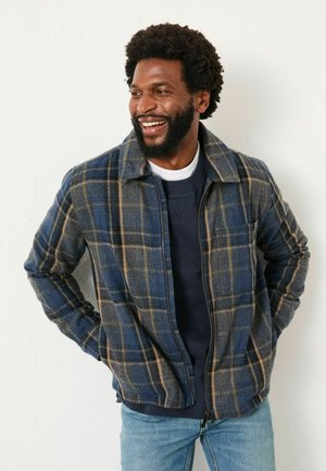 Plaid flannel jacket in blue, brown, and beige, features a collar and zip closure. Underneath, a navy sweatshirt is visible.