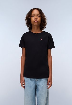 Schwarzes Baumwoll-T-Shirt mit Rundhalsausschnitt und kurzen Ärmeln, das ein kleines rotes und weißes Logo auf der Brust hat. Getragen mit hellblauen weiten Jeans.