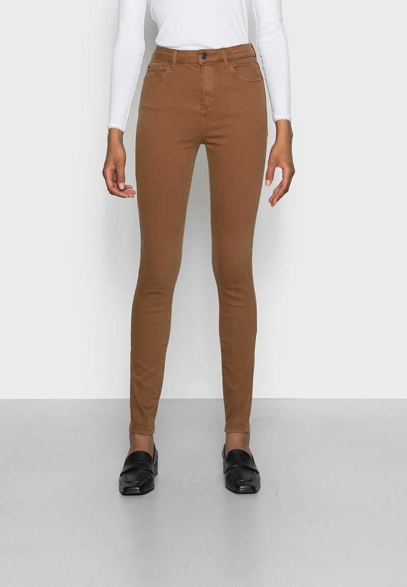 Pantalon slim marron porté avec un haut blanc à manches longues et des mocassins noirs, debout devant un fond clair uni.