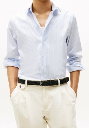 Homme portant une chemise à rayures bleu clair avec les manches retroussées, rentrée dans un pantalon blanc avec une ceinture noire tressée, les mains dans les poches.