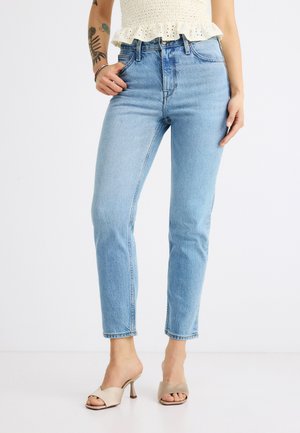 Personne portant un jean droit taille haute bleu clair, un top blanc sans manches, des sandales à talons beiges et des tatouages visibles sur les bras.