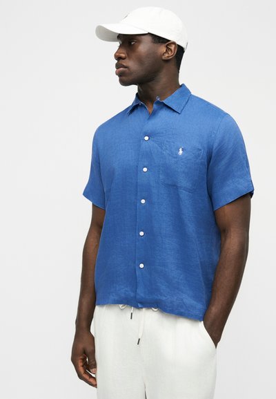 Vīrietis, kurš valkā zilu īss piedurkņu krekliņu ar krūšu kabatu un baltas bikses, skatās pa kreisi, valkā baltu cepuri.