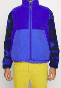 Chaqueta de forro polar morada con cierre de cremallera, mangas negras con un patrón en contraste y dos bolsillos frontales. Lleva puesta sobre pantalones de pana amarillos.