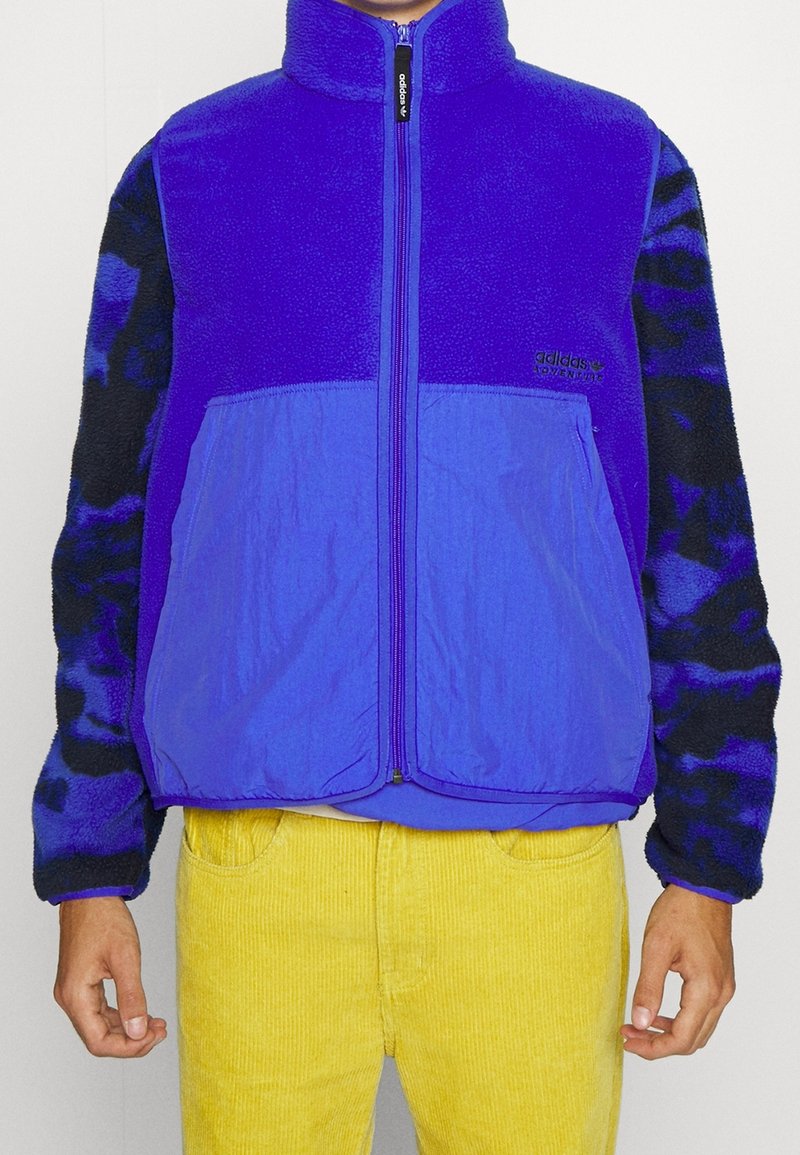 Chaqueta de forro polar morada con cierre de cremallera, mangas negras con un patrón en contraste y dos bolsillos frontales. Lleva puesta sobre pantalones de pana amarillos.