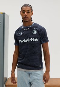 Camisola desportiva azul-marinho com padrão geométrico, mangas curtas, detalhes azuis e um emblema circular. Inclui a marca "MediaMarkt".