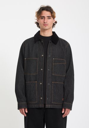 Joven con cabello castaño ondulado que lleva una chaqueta negra de rayas finas con bolsillos delanteros y cierre de botones, sobre un fondo blanco liso.