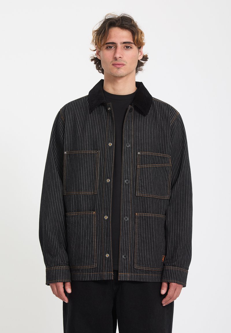 Jeune homme aux cheveux bruns ondulés portant une veste noire à fines rayures avec poches avant et fermeture par boutons sur un fond blanc uni.