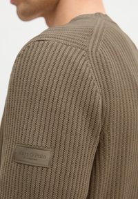 Gros plan sur un homme portant un pull en tricot côtelé taupe avec un patch rectangulaire Marc O’Polo sur la partie supérieure de la manche.