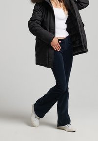 Černá prošívaná bunda s kapucí lemovanou kožešinou, bílý tílko a tmavě modré kordurové rozšířené kalhoty. Bílá teniska doplňuje outfit.