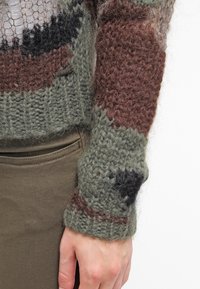 Neulepaita, jossa on teksturoitu, monivärinen kuvio vihreässä, ruskeassa ja mustassa, varustettuna ribattu hihansuu ja näkyvä brändilippu.