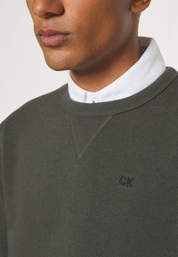 Olivengrønn sweatshirt med rund hals og ribbede mansjetter, som har en subtil brodert "CK" logo på brystet. Materialet er en bomullsblanding.