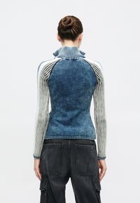 Kvinna med håret uppsatt i en knut, klädd i en blå- och vitrevärmad tröja med hög krage och svarta cargobyxor, vänd bort från kameran mot en enfärgad bakgrund.