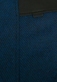 Blått texturerat tyg med ett fiskbensmönster och en svart läderdetalj. Inkluderar en liten, rektangulär varumärkesskylt märkt "G STAR."