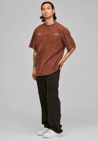 Camiseta de algodón marrón de gran tamaño con cuello de canalé y detalle de texto, combinada con pantalones cargo negros y zapatillas blancas, sobre un fondo gris.