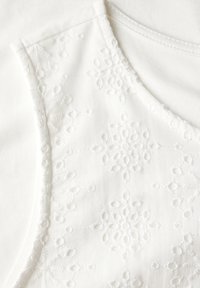 Vit tyg topp med ett texturerat broderat blommönster. Slät halsringning och axelsömmar, med subtil kontrast i tygdensitet.