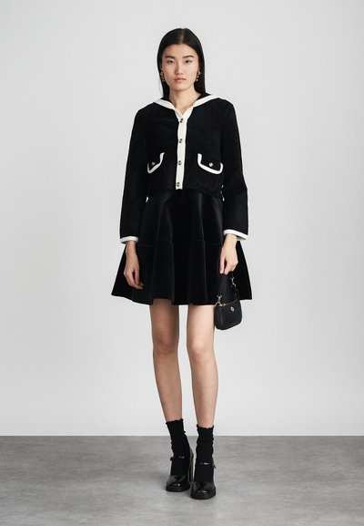 Schwarze, kurze Jacke mit weißem Kragen und Taschen-Details, kombiniert mit einem schwarzen Samtrock. Das Modell trägt schwarze Socken und klobige Schuhe.