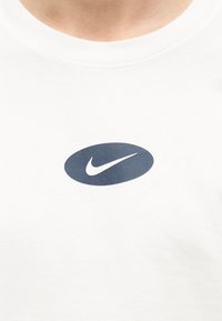 Λευκό πουκάμισο με ένα μικρό σκούρο οβάλ και το λευκό λογότυπο Nike Swoosh στο κέντρο του στήθους.