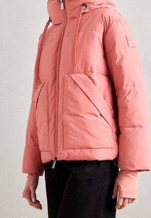 Žena má na sobě korálově růžovou prošívanou bundu s kapucí, předním zipem, velkými předními kapsami a manžetami s otvory na palce, v kombinaci s černými kalhotami.