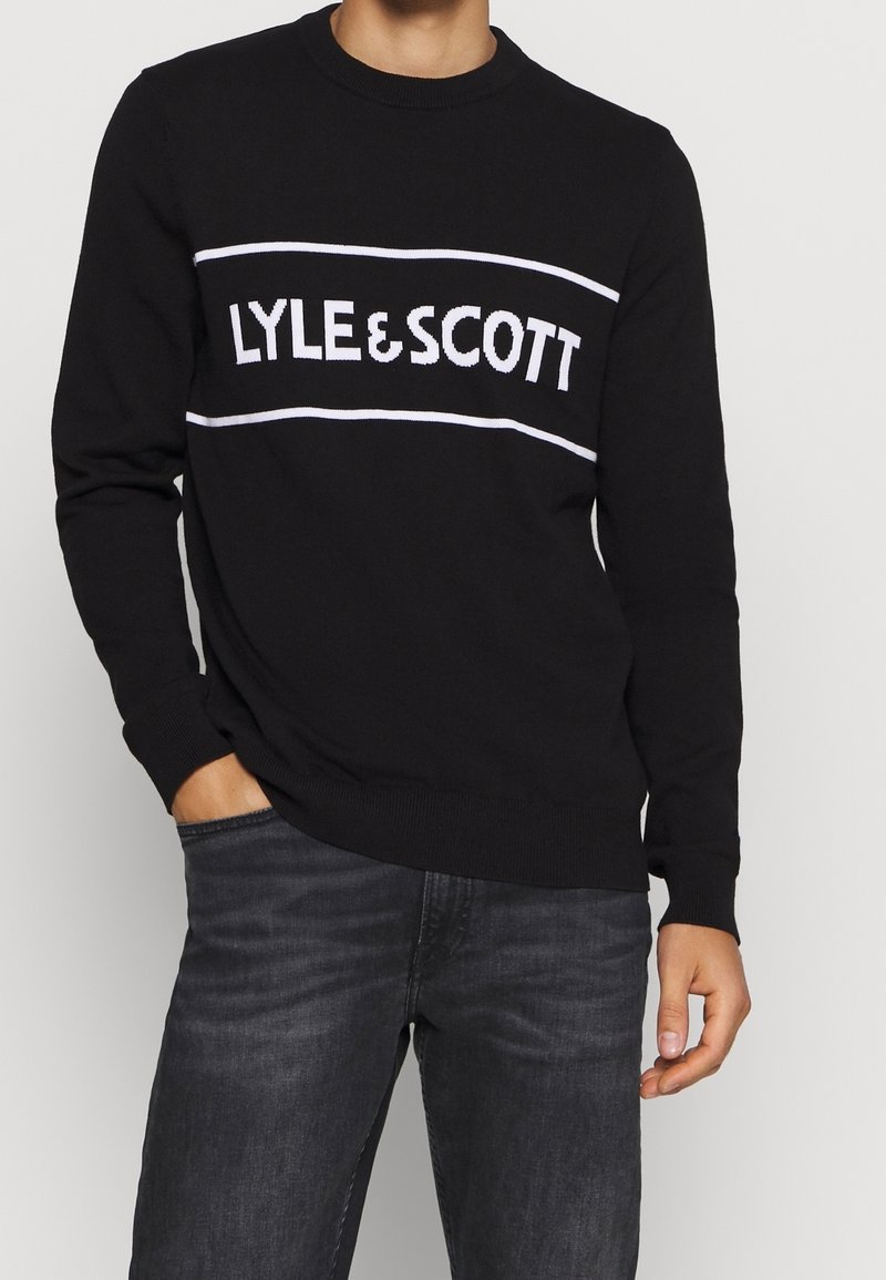 Pull noir avec un col ras du cou et des poignets côtelés comportant une bande horizontale blanche affichant "LYLE & SCOTT" en texte gras.