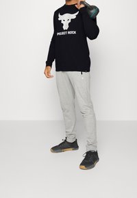 Chemise noire à manches longues avec logo de taureau, pantalon de sport gris et chaussures de sport noires avec semelles en caoutchouc. Tient un kettlebell.