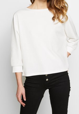 Femme portant un haut blanc ample à manches longues avec des poignets côtelés et un pantalon noir taille haute avec deux boutons visibles.