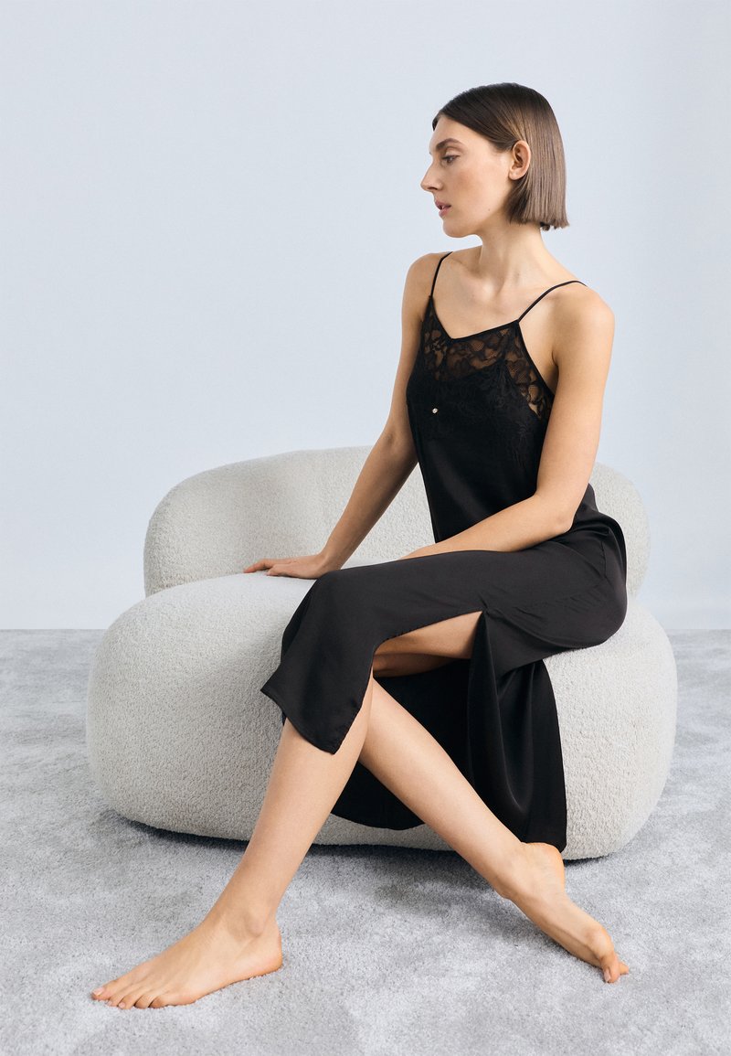 Rochie din satin negru cu detalii din dantelă, cu bretele subțiri și o fustă cu șliț lateral, purtată de un model așezat pe un scaun texturat, de culoare deschisă.