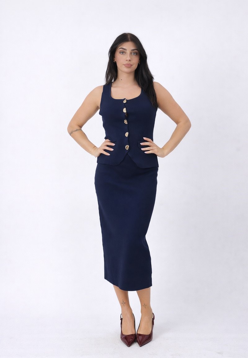 Vrouw staand met handen in de zij, draagt een marineblauwe vest met knopen en een bijpassende lange rok, donker haar en bordeauxrode spitse hakken.