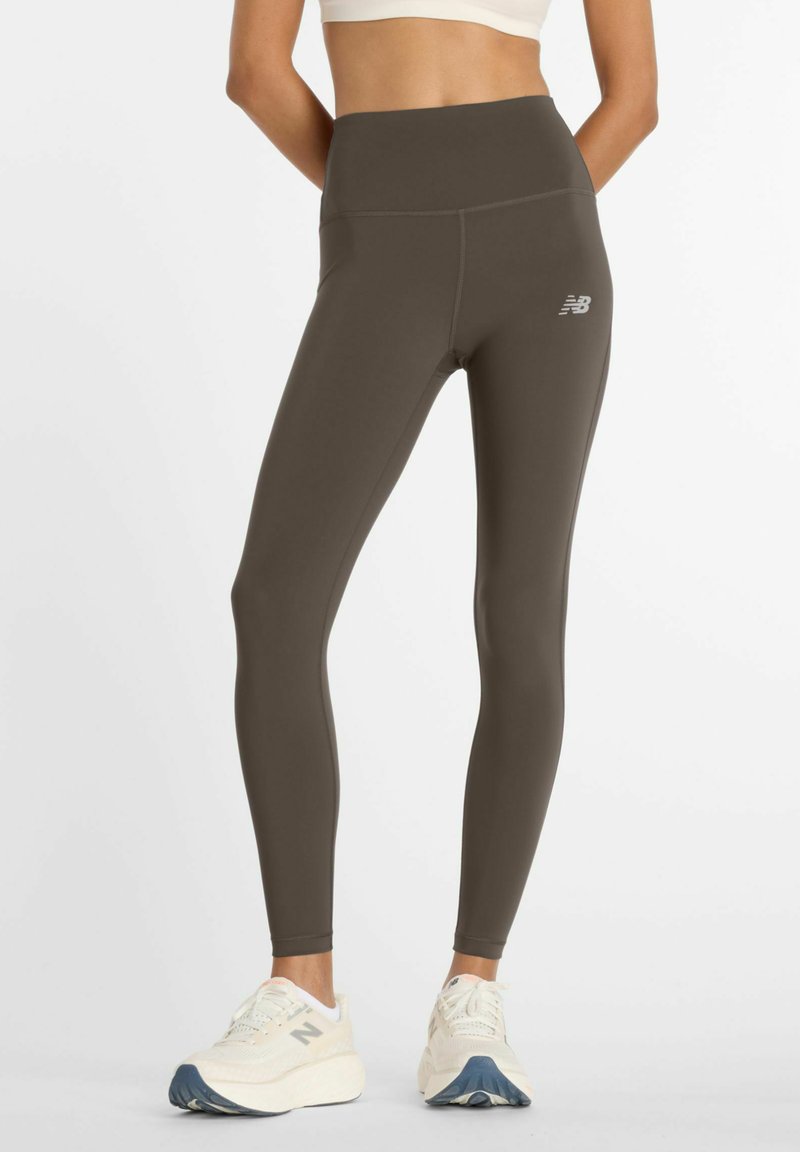 Femme portant des leggings New Balance taille haute marron foncé et des baskets blanches New Balance, debout devant un fond blanc uni.