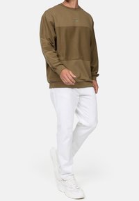Man wearing olive green color-block sweatshirt, white slim-fit pants, and white chunky sneakers, walking against neutral background.