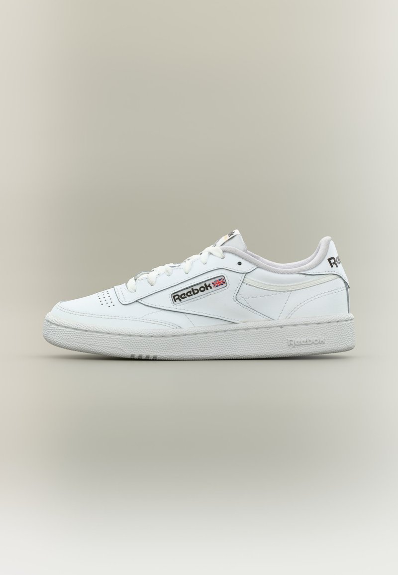 Zapatilla blanca de caña baja Reebok con cordones en la parte frontal, puntera perforada, suela texturizada y logo de Reebok con la bandera británica en el lateral y el talón.